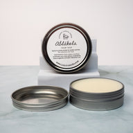 An open tin of cream colored balm next to a tin lid with a label that says Lakota Made Ablakela calm balm. The tins are on a gray marble surface in front of a white background. The ingredients are shea butter, beeswax, sunflower oil, rose petals, chamomile, hops, mugwort, and lavender essential oil.