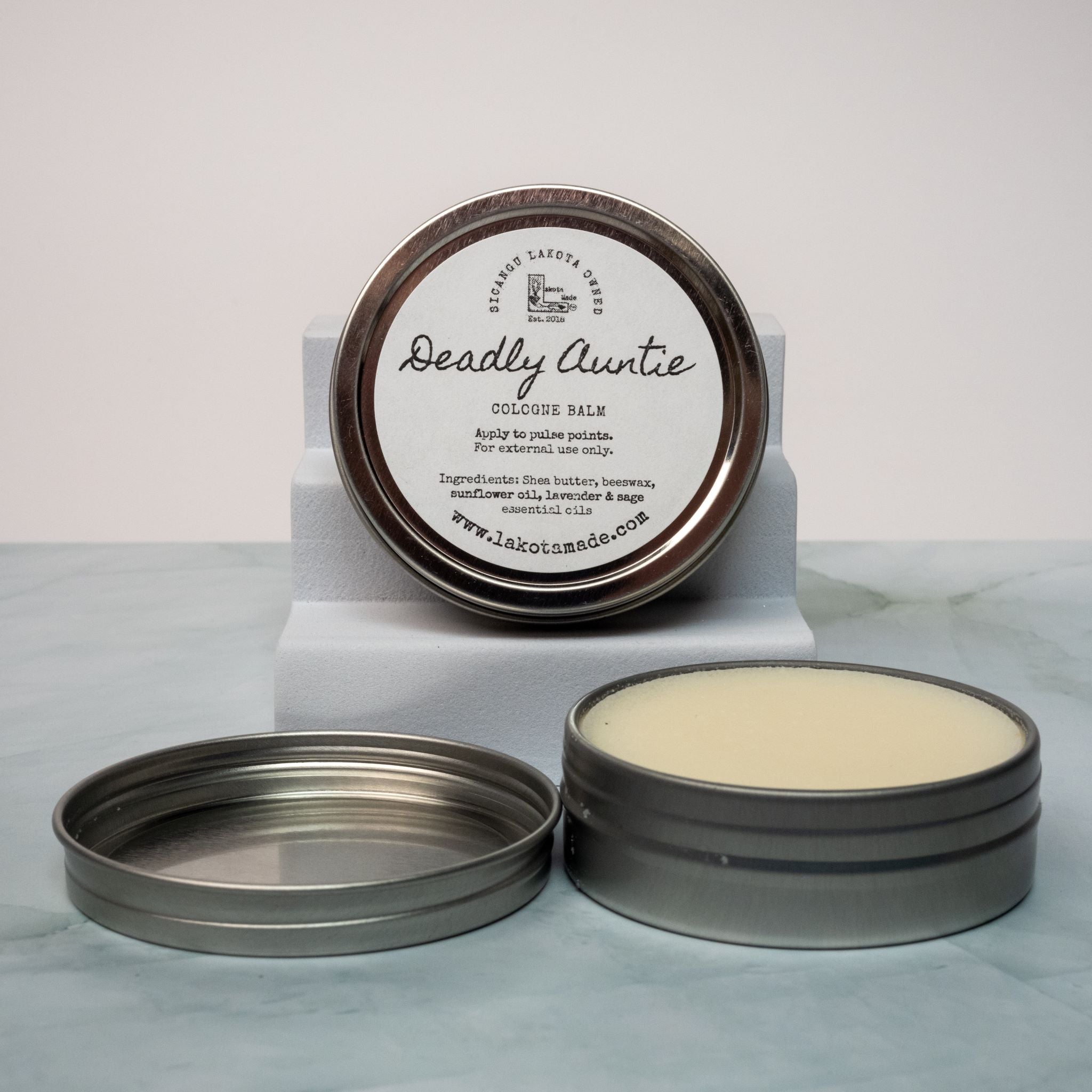 An open tin of cream colored balm next to a tin lid with a label that says Lakota Made Deadly Auntie balm. The tins are on a gray marble surface in front of a white background. The ingredients are shea butter, beeswax, sunflower oil, sage and lavender essential oil.