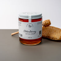 A round jar of elderberry infused honey positioned on a warm gray surface with a white background. A white label on the jar says Lakota Made Elderberry Infused Honey 8 fl. oz. Behind the glass jar is a piece of honeycomb and a honey dipper.