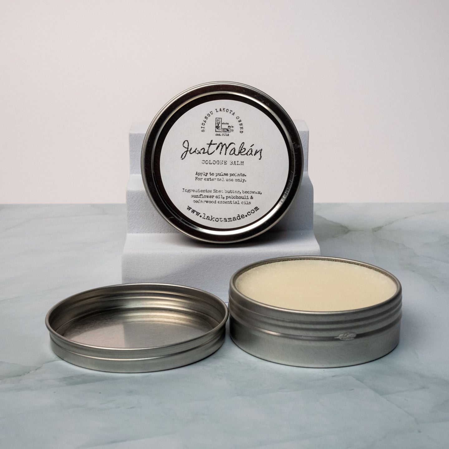 An open tin of cream colored balm next to a tin lid with a label that says Lakota Made Just Wakan cologne balm. The tins are on a gray marble surface in front of a white background. The ingredients are shea butter, beeswax, sunflower oil, patchouli and cedarwood essential oil.