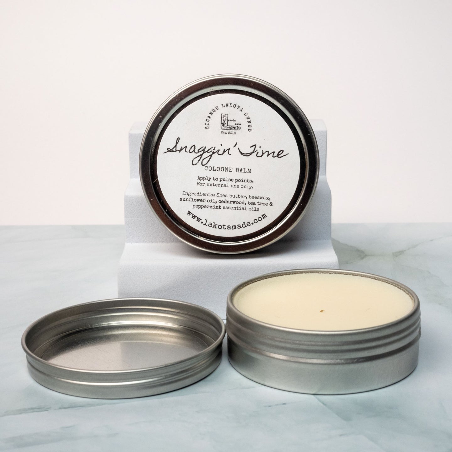 An open tin of cream colored balm next to a tin lid with a label that says Lakota Made Snaggin' Time cologne balm. The tins are on a gray marble surface in front of a white background. The ingredients are shea butter, beeswax, sunflower oil, cedarwood, tea tree, and peppermint essential oil.