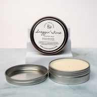 An open tin of cream colored balm next to a tin lid with a label that says Lakota Made Snaggin' Time cologne balm. The tins are on a gray marble surface in front of a white background. The ingredients are shea butter, beeswax, sunflower oil, cedarwood, tea tree, and peppermint essential oil.