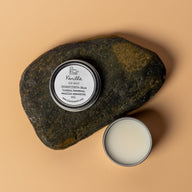 A round aluminum tin containing a cream colored lip balm. The lid says vanilla lip balm ingredients shea butter beeswax vanilla essential oil. The lid is positioned on a dark brown rock on a cream orange background.