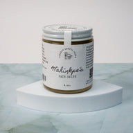 A round glass container containing a yellow cream medicinal salve. The glass has a white label that says Lakota Made Wahiŋkpe Pain Salve 4 oz. ingredients Beeswax, Lavender, Plantain, Comfrey, Yarrow, Calendula, Chamomile, Arnica Flower, Ginger Root, Peppermint, Magnesium & Sunflower Oil. The glass jar is positioned on a wedge shaped white riser on a blue gray marble surface with a white background.