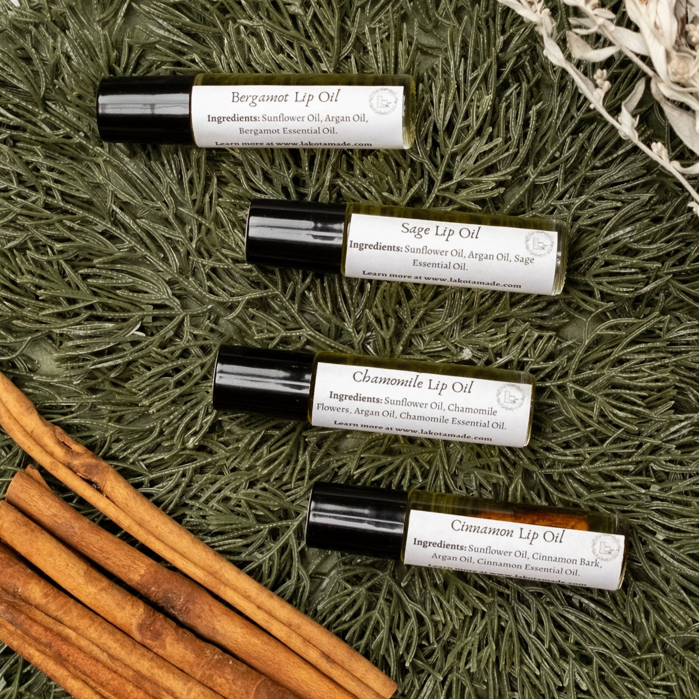 Four clear glass roller bottles with black lids and white labels that say Lakota Made Bergamot Lip Oil, Sage Lip Oil, Chamomile Lip Oil, and Cinnamon Lip Oil on a pine needle background with dried sage and cinnamon sticks.
