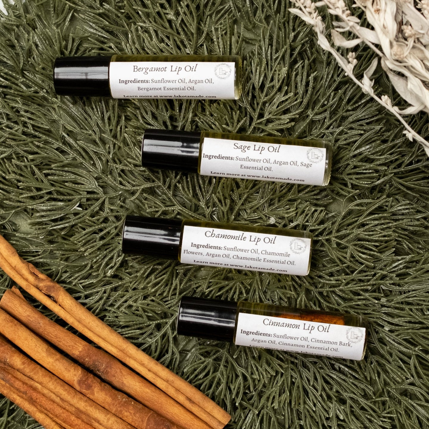 Collection photo for Cinnamon Lip Oil. Four clear glass roller bottles with black lids and white labels that say Lakota Made Bergamot Lip Oil, Sage Lip Oil, Chamomile Lip Oil, and Cinnamon Lip Oil on a pine needle background with dried sage and cinnamon sticks.