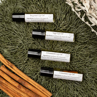Collection photo for Cinnamon Lip Oil. Four clear glass roller bottles with black lids and white labels that say Lakota Made Bergamot Lip Oil, Sage Lip Oil, Chamomile Lip Oil, and Cinnamon Lip Oil on a pine needle background with dried sage and cinnamon sticks.