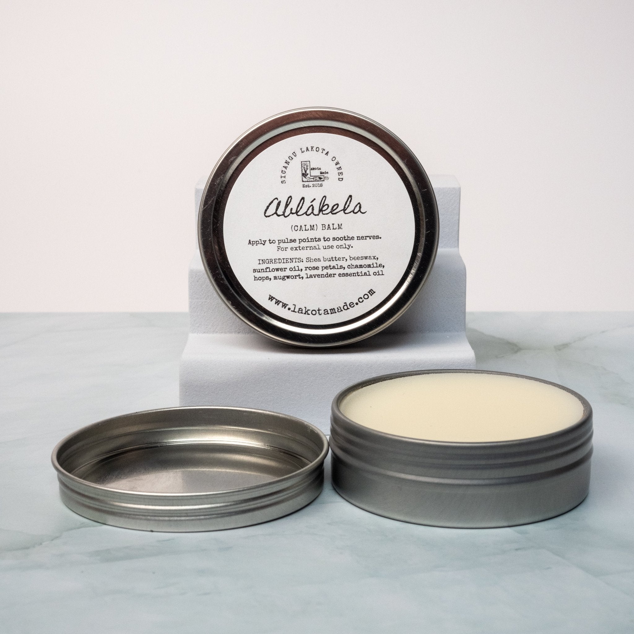 An open tin of cream colored balm next to a tin lid with a label that says Lakota Made Ablakela calm balm. The tins are on a gray marble surface in front of a white background. The ingredients are shea butter, beeswax, sunflower oil, rose petals, chamomile, hops, mugwort, and lavender essential oil. 