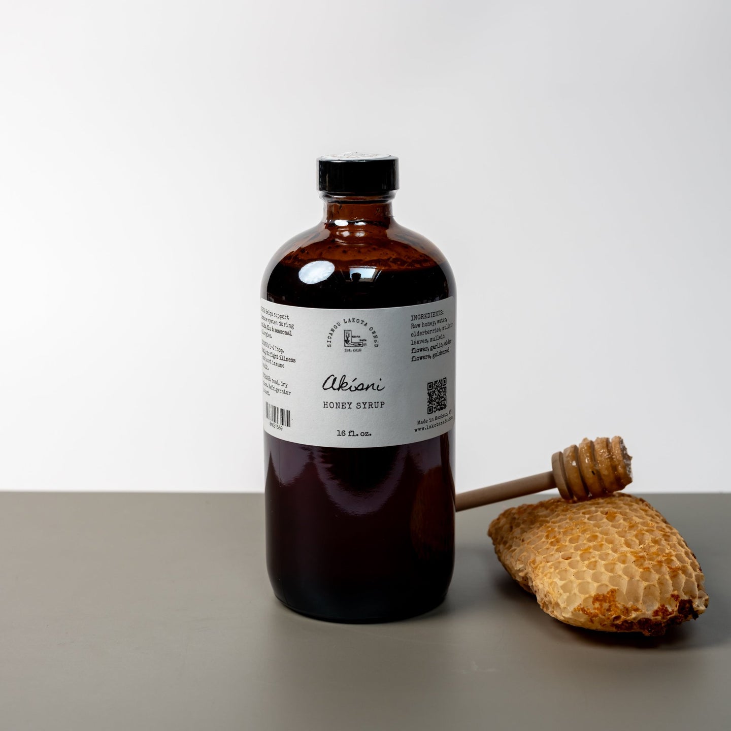 A large brown bottle with a white label that says Lakota Made Akisni Honey Syrup 16 fl oz. Ingredients are raw honey, elderberries, mullein flowers, garlic, goldenrod. There is a honey comb and dipper next to the bottle. It is resting on a gray table top in front of a white background.