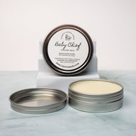 An open aluminum tin of cologne balm with a white label that says Lakota Made Baby Chief Cologne Balm. Ingredients: Shea butter, beeswax, sunflower oil, cedarwood & Sage essential oils. The tin in on a white marble surface in front of a white background.