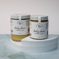 Two glass jars of Lakota Made Baby Love Healing Salve with white lids and labels. Ingredients are sunflower oil, shea butter, beeswax, plantain, yarrow, white willow, and jewelweed. The jars are on a white pedestal stand on a white marble surface with white background.