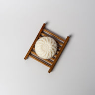 A white bar of soap shaped like a round flower on a bamboo ladder style soap dish on a white background.