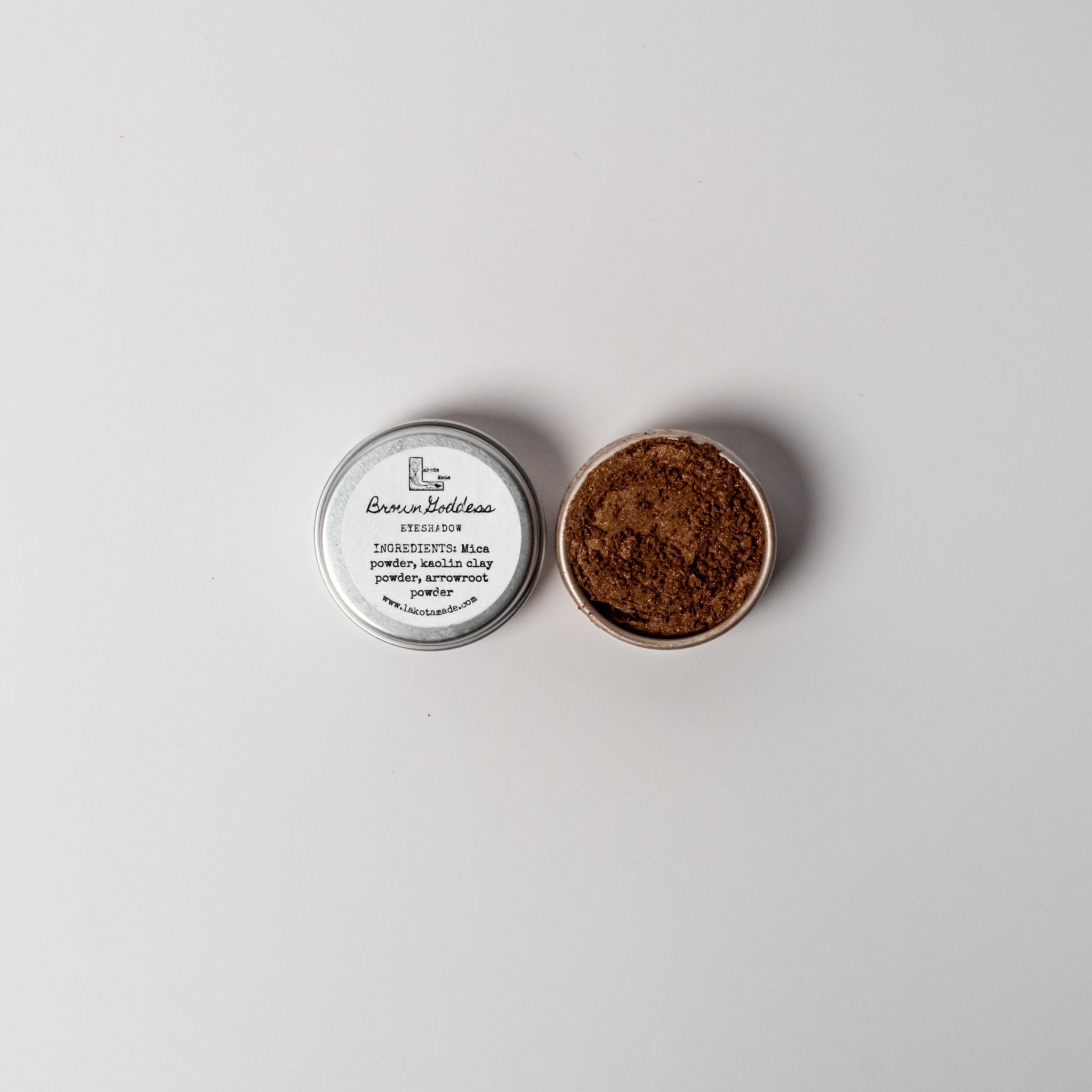 An open aluminum tin or rich brown powder on a white background with a white label that says Lakota Made Brown Goddess Eyeshadow Ingredients mica powder, kaolin clay powder, arrowroot powder.