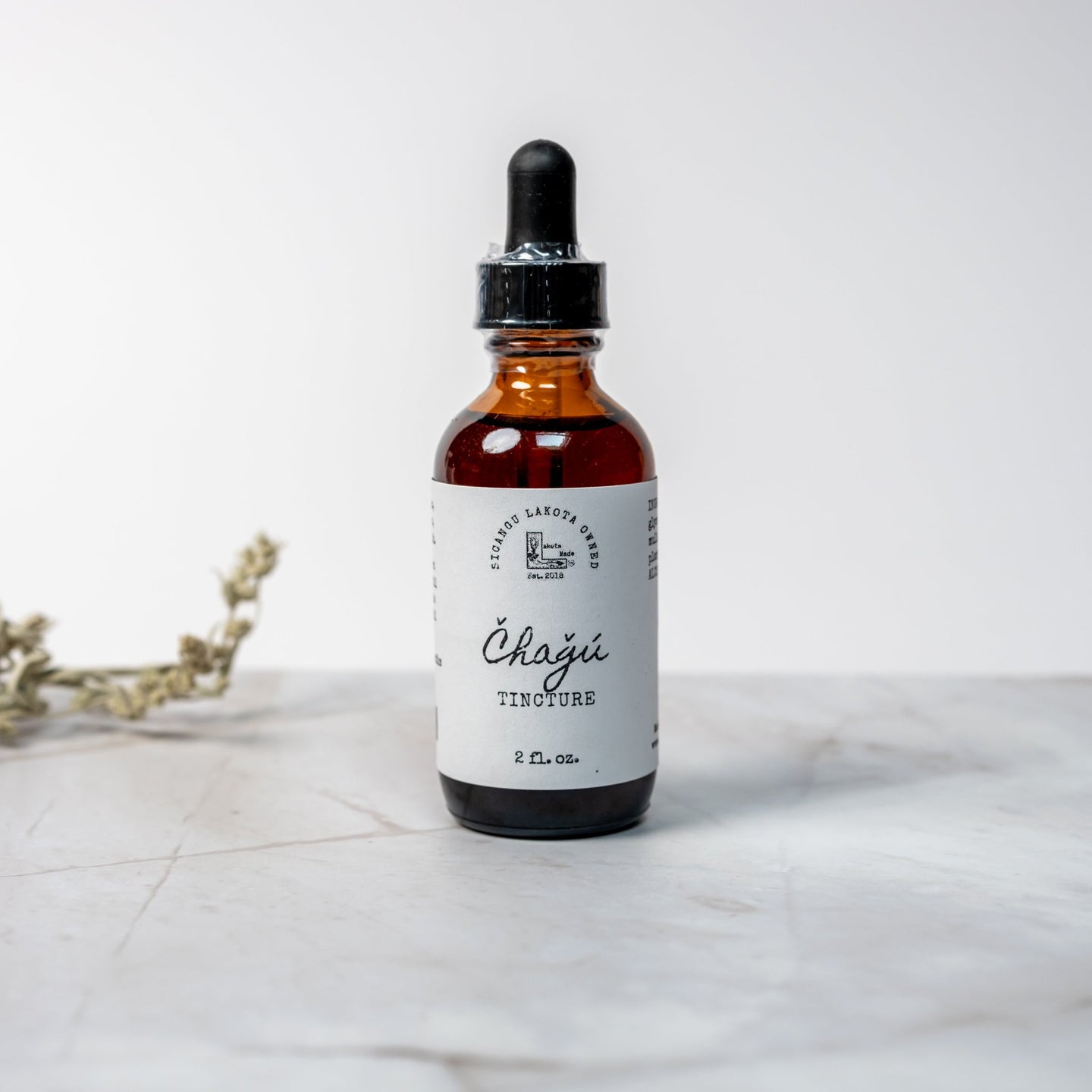 A brown dropper bottle with a white label that says Lakota Made Chagu Tincture 2 fl. oz. The bottle is on a white marble surface with a white background and dried plant.