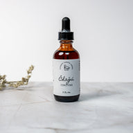 A brown dropper bottle with a white label that says Lakota Made Chagu Tincture 2 fl. oz. The bottle is on a white marble surface with a white background and dried plant.