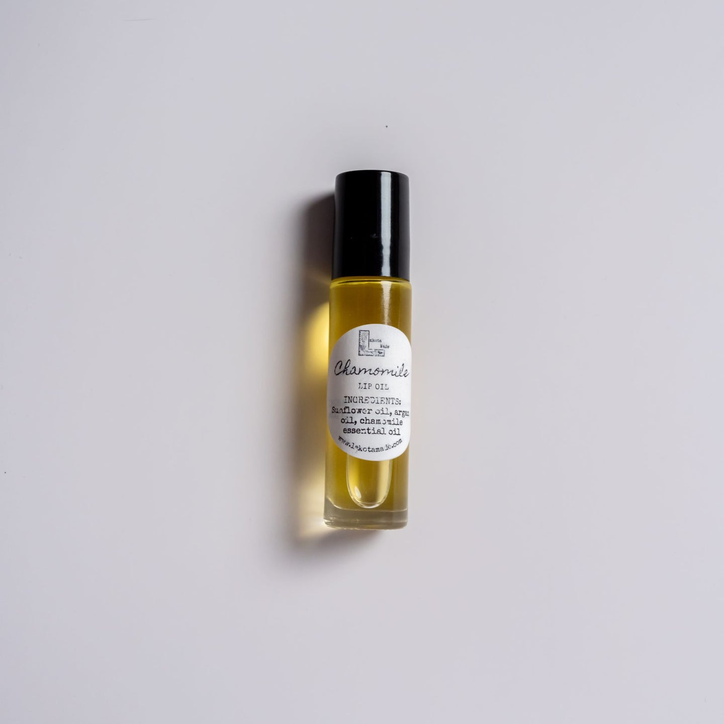 A clear glass roller bottle with a black lid and white label that says Lakota Made Chamomile Lip Oil ingredients sunflower oil, argan oil, chamomile essential oil.