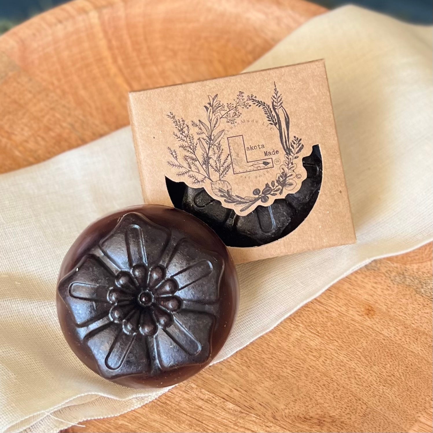 A close up of a round flower shaped brown soap bar with a second bar in a brown cardboard package. The package says Lakota Made. The products are on a white cloth in a wooden dish.