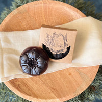 A round flower shaped dark brown soap bar with a second bar in a brown cardboard package. The package says Lakota Made. The products are on a cloth in a wooden dish surrounded by pine needles.