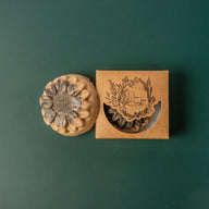 A round flower shaped yellow brown soap bar with a second bar in a brown cardboard package. The package says Lakota Made. The products are on a dark green background.