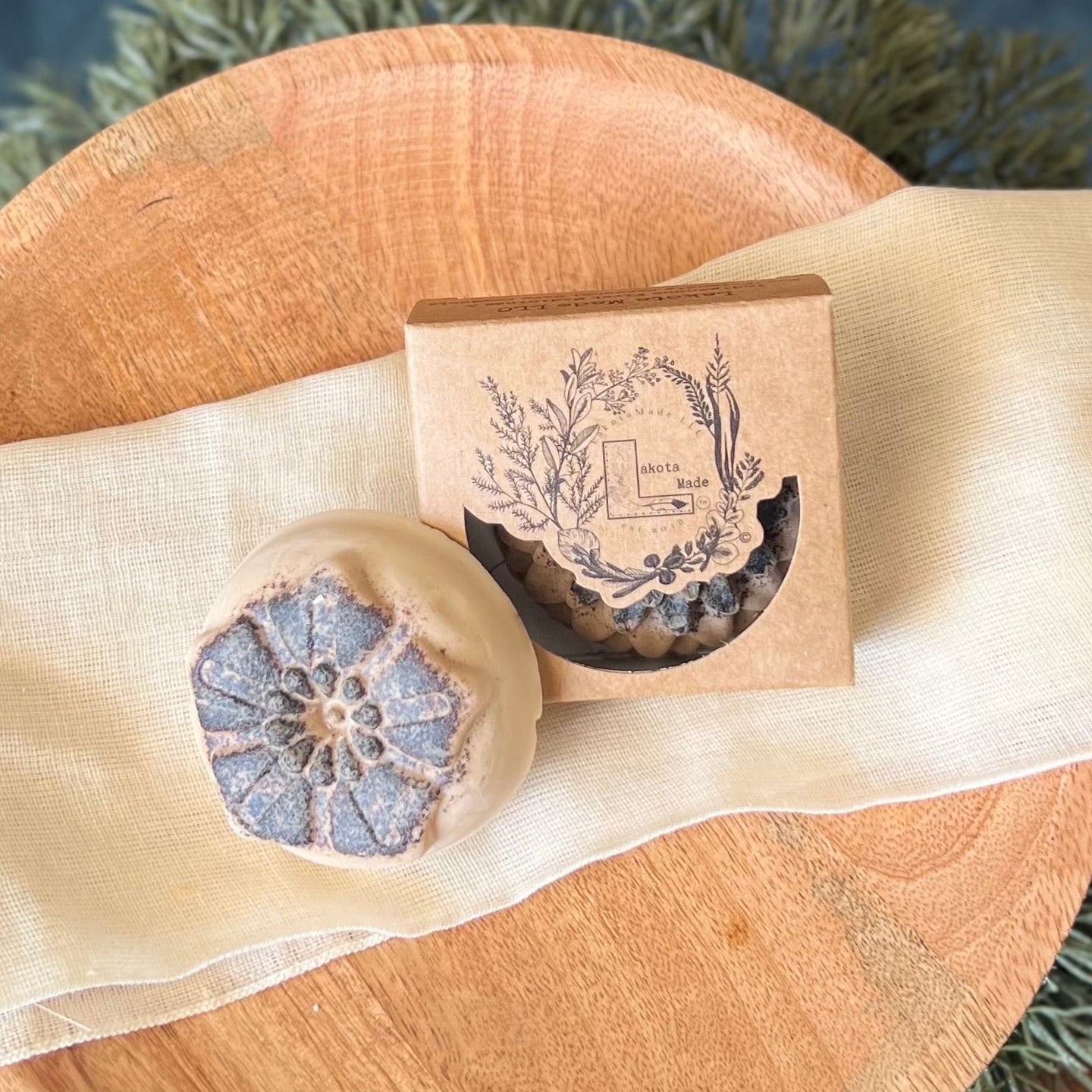 A round flower shaped cream and brown soap bar with a second bar in a brown cardboard package. The package says Lakota Made. The products are on a white cloth in a wooden dish surrounded by pine needles.