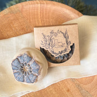 A closeup of a round flower shaped cream and brown soap bar with a second bar in a brown cardboard package. The package says Lakota Made. The products are on a white cloth in a wooden dish.