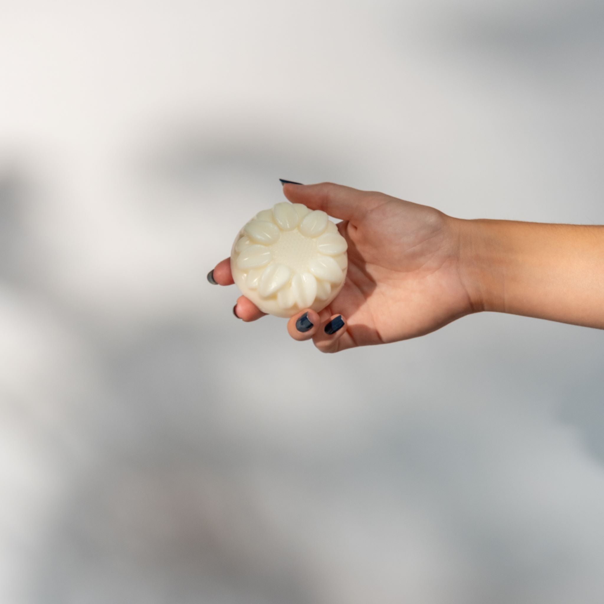 A woman's hand holding a round flower shaped cream colored soap bar on a gray background.