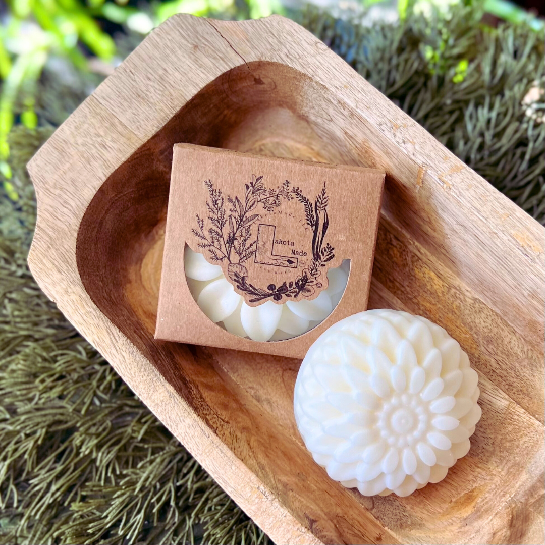 A round flower shaped cream colored soap bar with a second bar in a brown cardboard package. The package says Lakota Made. The products are in a wooden dish surrounded by pine needles.