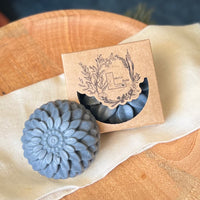 A close up of a round flower shaped gray colored soap bar with a second bar in a brown cardboard package. The package says Lakota Made. The products are on a cloth in a wooden dish surrounded by pine needles.