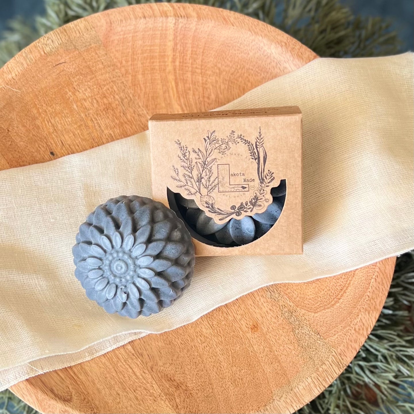 A round flower shaped gray colored soap bar with a second bar in a brown cardboard package. The package says Lakota Made. The products are on a cloth in a wooden dish surrounded by pine needles.