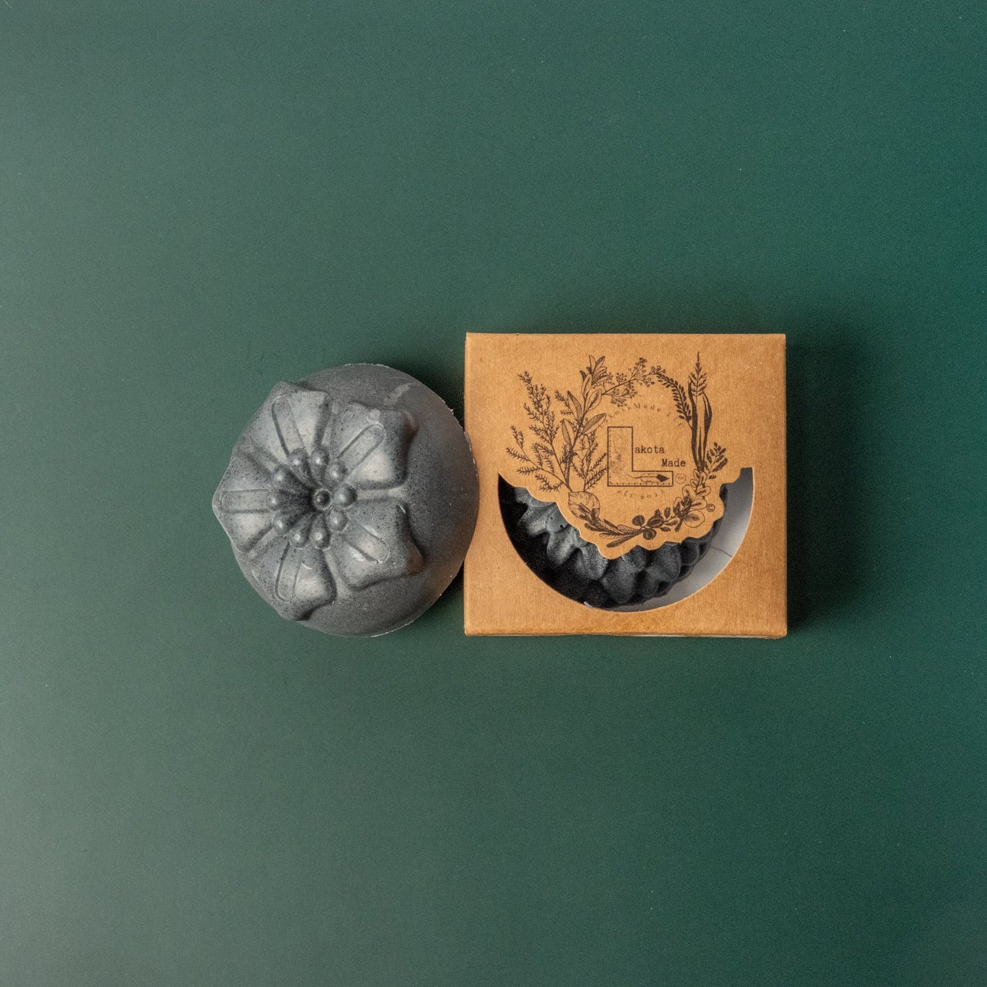 A round flower shaped gray soap bar with a second bar in a brown cardboard package. The package says Lakota Made. The products are on a dark green background.