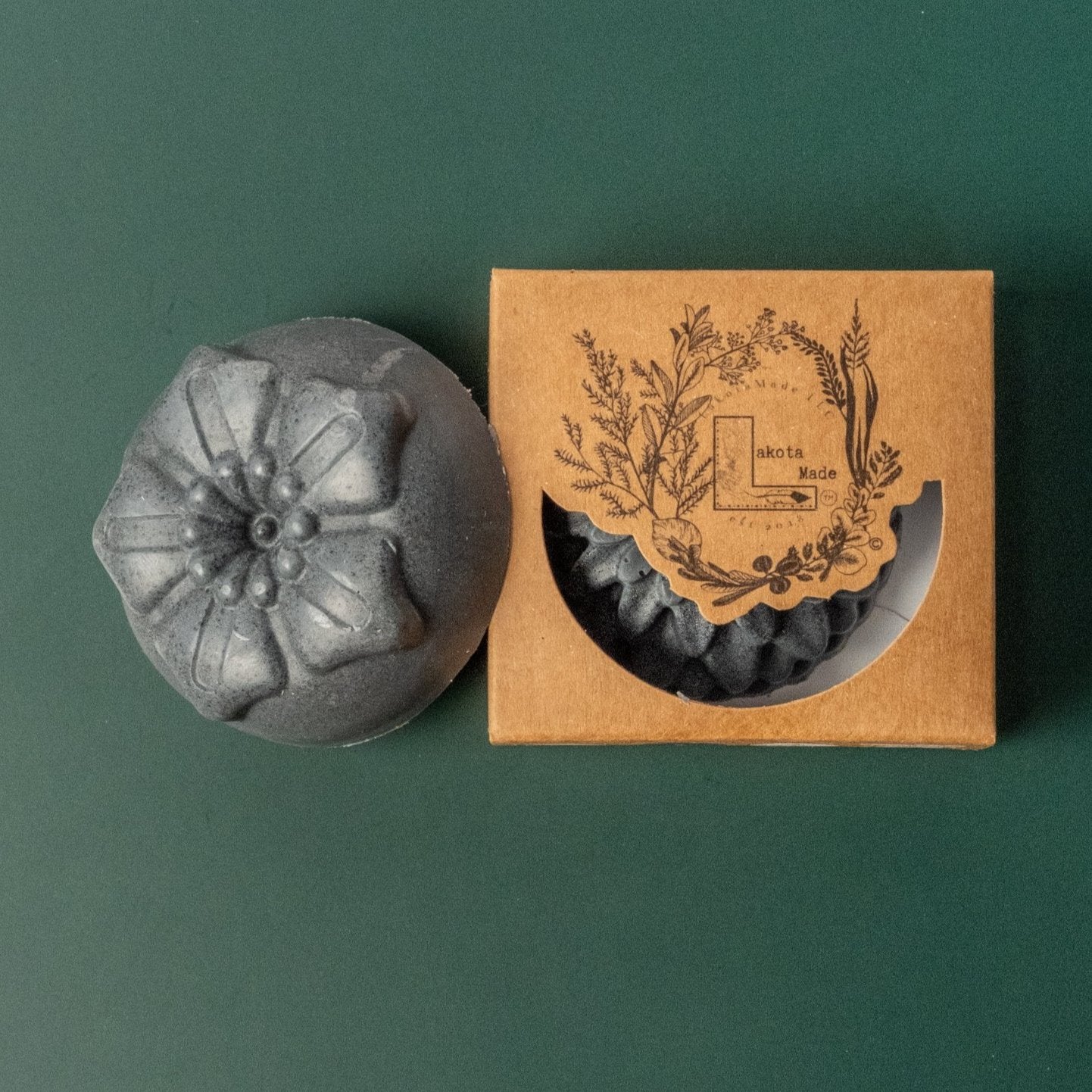 A round flower shaped gray soap bar with a second bar in a brown cardboard package. The package says Lakota Made. The products are on a dark green background.