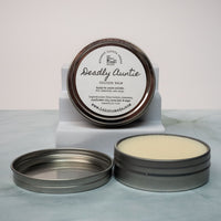 An open tin of cream colored balm next to a tin lid with a label that says Lakota Made Deadly Auntie balm. The tins are on a gray marble surface in front of a white background. The ingredients are shea butter, beeswax, sunflower oil, sage and lavender essential oil. 