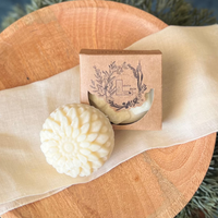 A round flower shaped cream colored soap bar with a second bar in a brown cardboard package. The package says Lakota Made. The products are on a cloth in a wooden dish surrounded by pine needles.