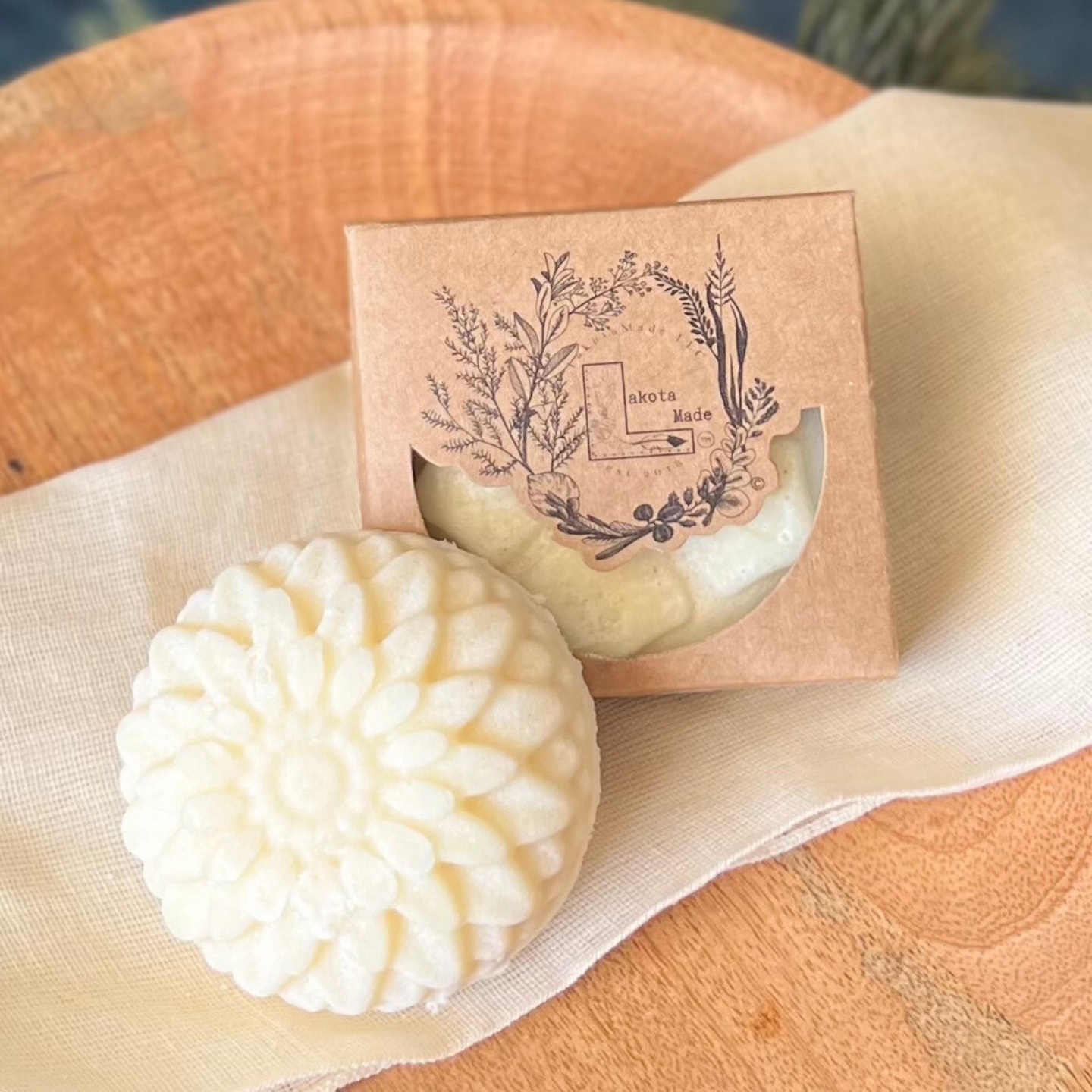 A closeup of a round flower shaped cream colored soap bar with a second bar in a brown cardboard package. The package says Lakota Made. The products are on a cloth in a wooden dish surrounded by pine needles.