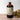 A large brown bottle with a white label that says Lakota Made Elderberry Chaga Tonic. There is a bundle of dried flowers next to the bottle. It is resting on a wooden table top in front of a white background.