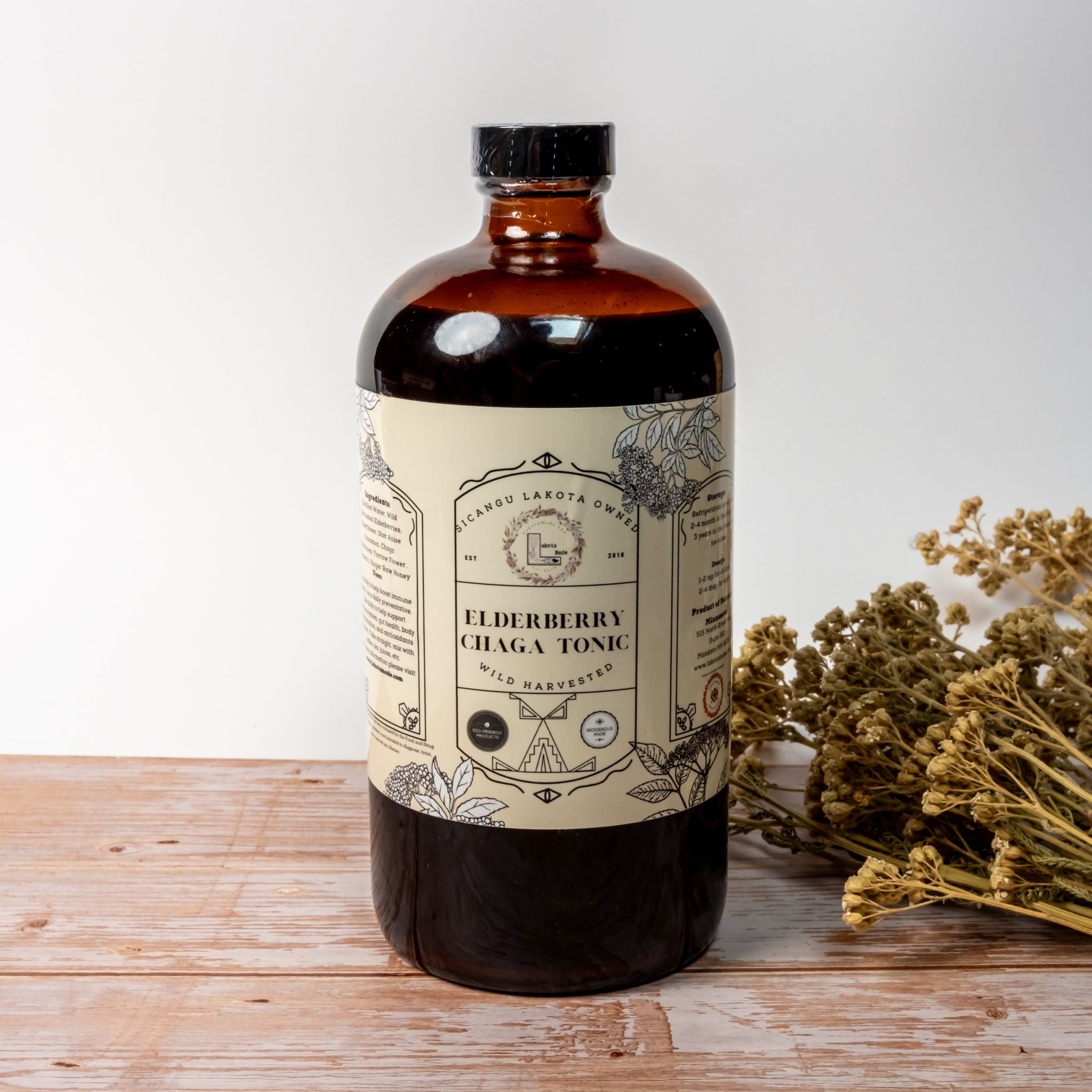 A large brown bottle with a white label that says Lakota Made Elderberry Chaga Tonic. There is a bundle of dried flowers next to the bottle. It is resting on a wooden table top in front of a white background.