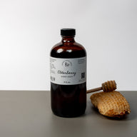 A large brown bottle with a white label that says Lakota Made Elderberry Honey Syrup 16 fl oz. Ingredients are raw honey, and elderberries. There is a honey comb and dipper next to the bottle. It is resting on a gray table top in front of a white background.