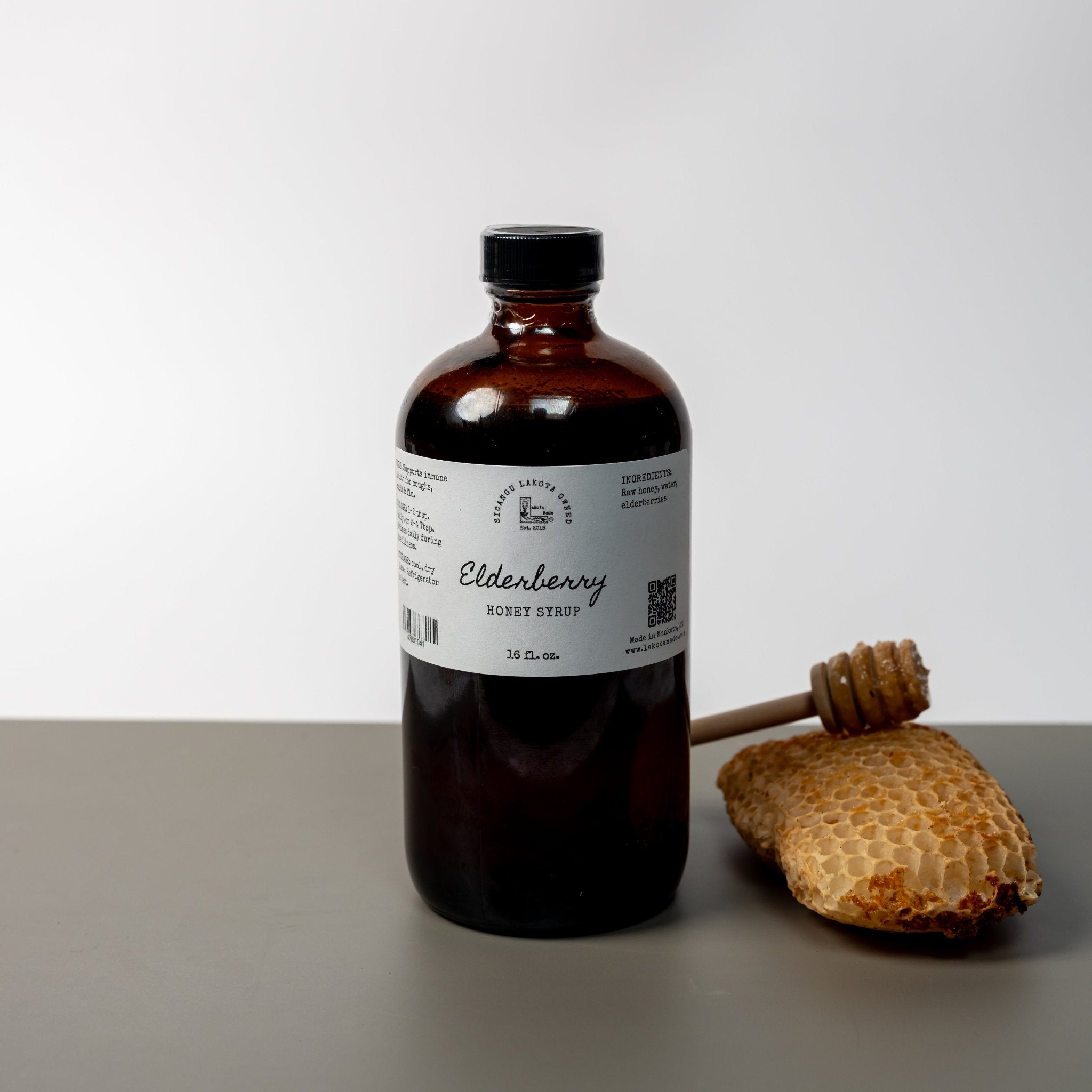 A large brown bottle with a white label that says Lakota Made Elderberry Honey Syrup 16 fl oz. Ingredients are raw honey, and elderberries. There is a honey comb and dipper next to the bottle. It is resting on a gray table top in front of a white background.