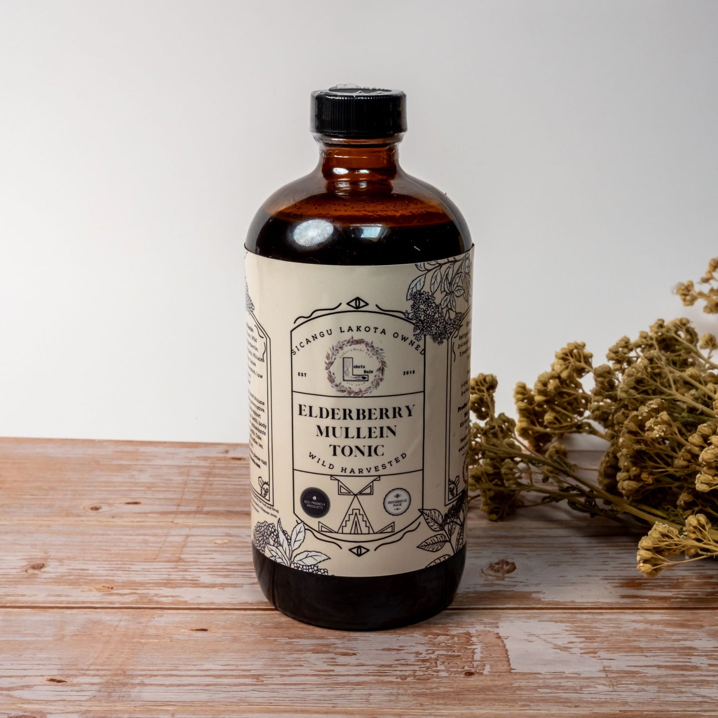 A large brown bottle with a white label that says Lakota Made Elderberry Mullein Tonic. There is a bundle of dried flowers next to the bottle. It is resting on a wooden table top in front of a white background.