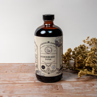 A large brown bottle with a white label that says Lakota Made Elderberry Chaga Tonic. There is a bundle of dried flowers next to the bottle. It is resting on a wooden table top in front of a white background.