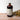 A large brown bottle with a white label that says Lakota Made Elderberry Tonic. There is a bundle of dried flowers next to the bottle. It is resting on a wooden table top in front of a white background.