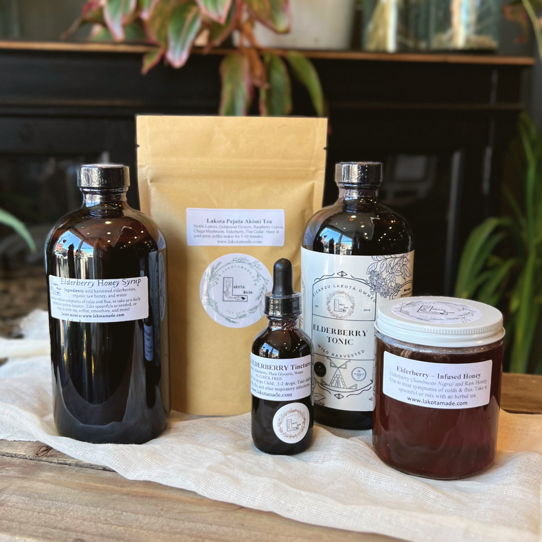 A collection of Lakota Made products including Elderberry Honey Syrup, Lakota Pejuta Akisni Tea, Elderberry Tincture, Elderberry Tonic, and Elderberry-Infused Honey. The bottles, jars, and bag are on a white cloth on a wooden table top in front of plants.