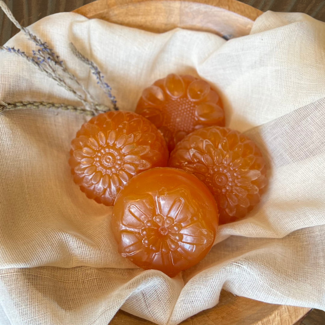 Four flower shaped translucent amber colored soap bars on a cloth in a wooden dish with dried lavender sprigs.