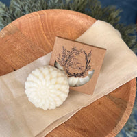 A round flower shaped cream colored soap bar with a second bar in a brown cardboard package. The package says Lakota Made. The products are on a cloth in a wooden dish surrounded by pine needles.