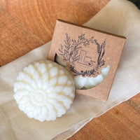 A close up of a round flower shaped cream colored soap bar with a second bar in a brown cardboard package. The package says Lakota Made. The products are on a cloth in a wooden dish surrounded by pine needles.