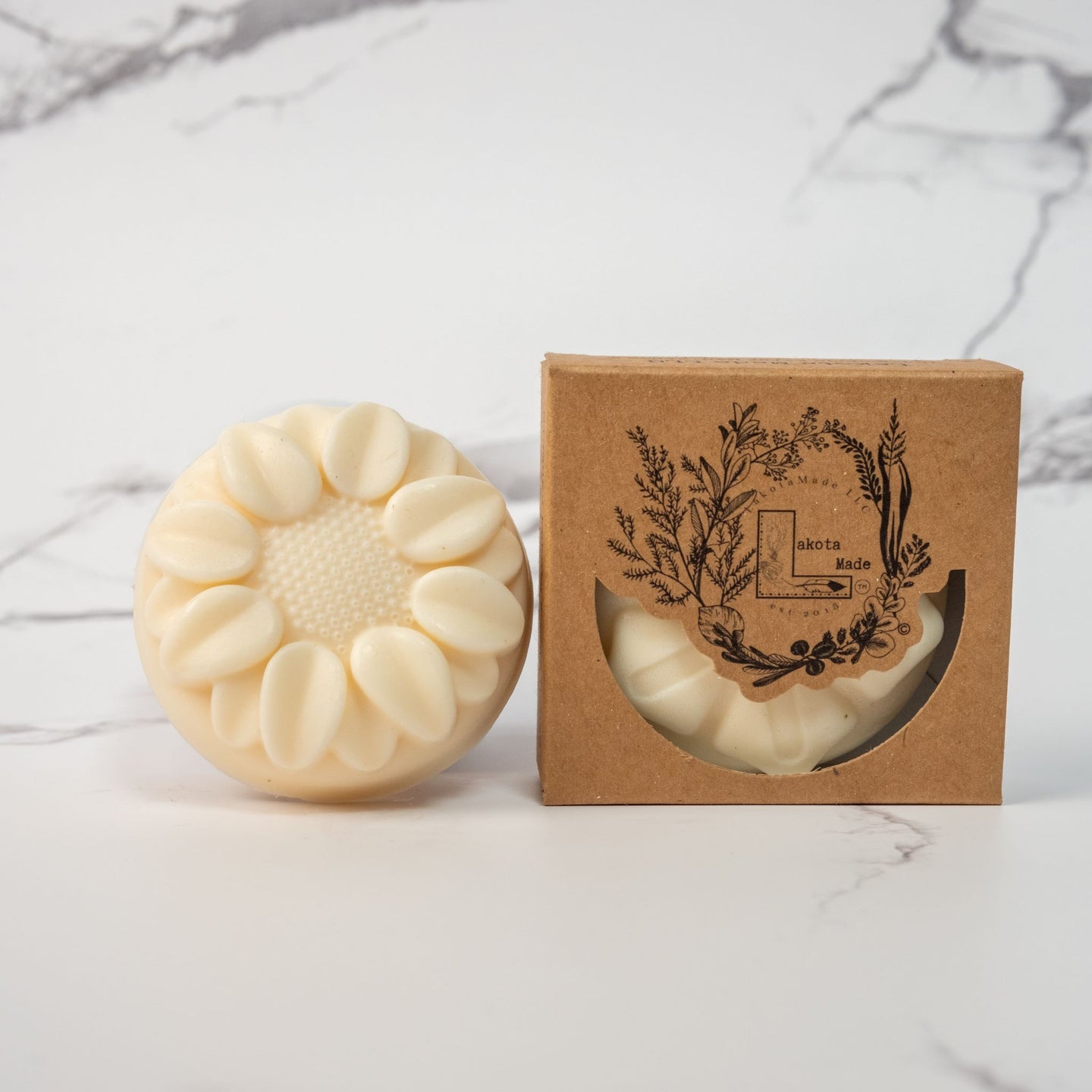 A close up of a round flower shaped cream colored soap bar with a second bar in a brown cardboard package. The package says Lakota Made. The products are on a white marble backround.