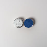 An open aluminum tin of vibrant blue powder on a white background with a white label that says Lakota Made  Hazatho Blue Eyeshadow Ingredients blue spirulina powder and arrowroot powder.