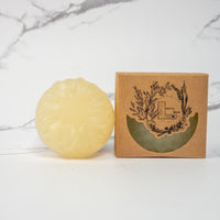 A round flower shaped translucent yellow colored soap bar with a second bar in a brown cardboard package. The package says Lakota Made. The products are on a white marble background.