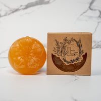 A round flower shaped translucent orange colored soap bar with a second bar in a brown cardboard package. The package says Lakota Made. The products are on a white marble background.