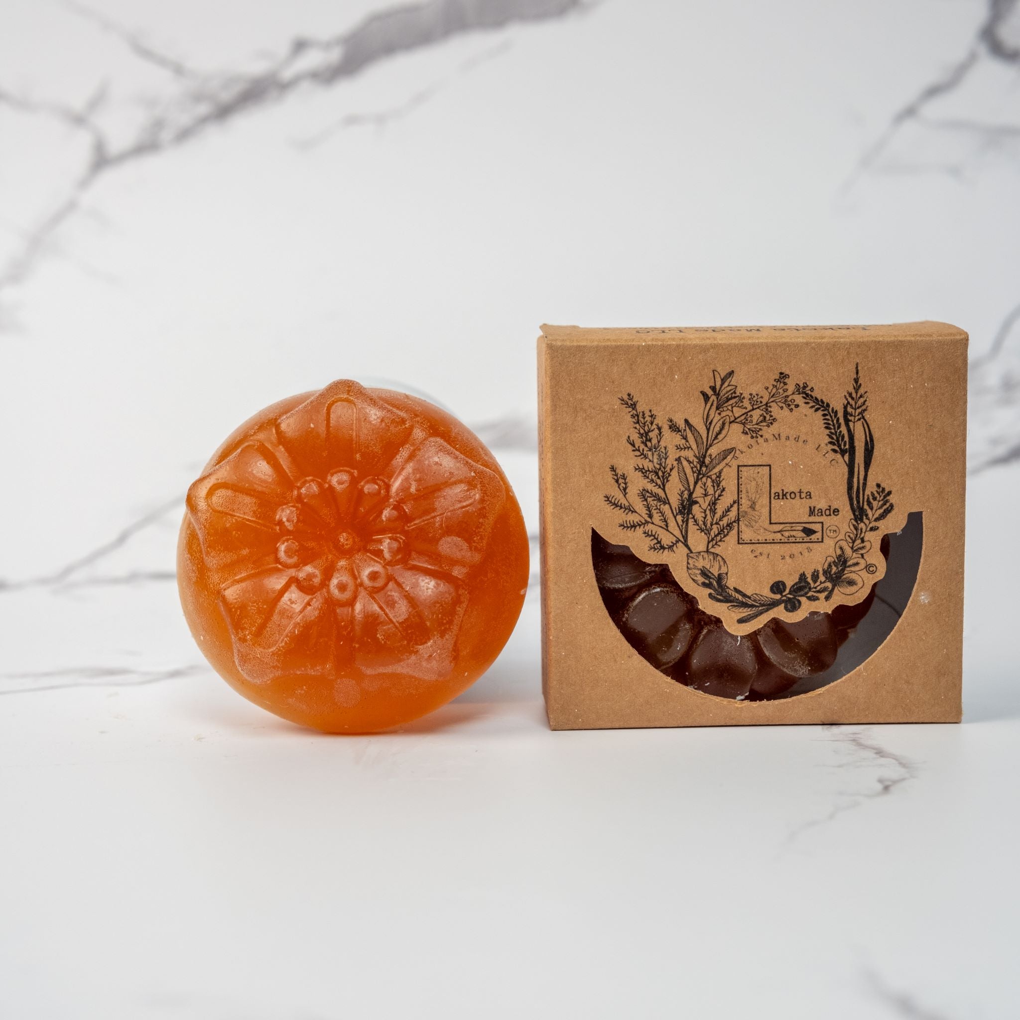 A round flower shaped translucent red orange colored soap bar with a second bar in a brown cardboard package. The package says Lakota Made. The products are on a white marble background.